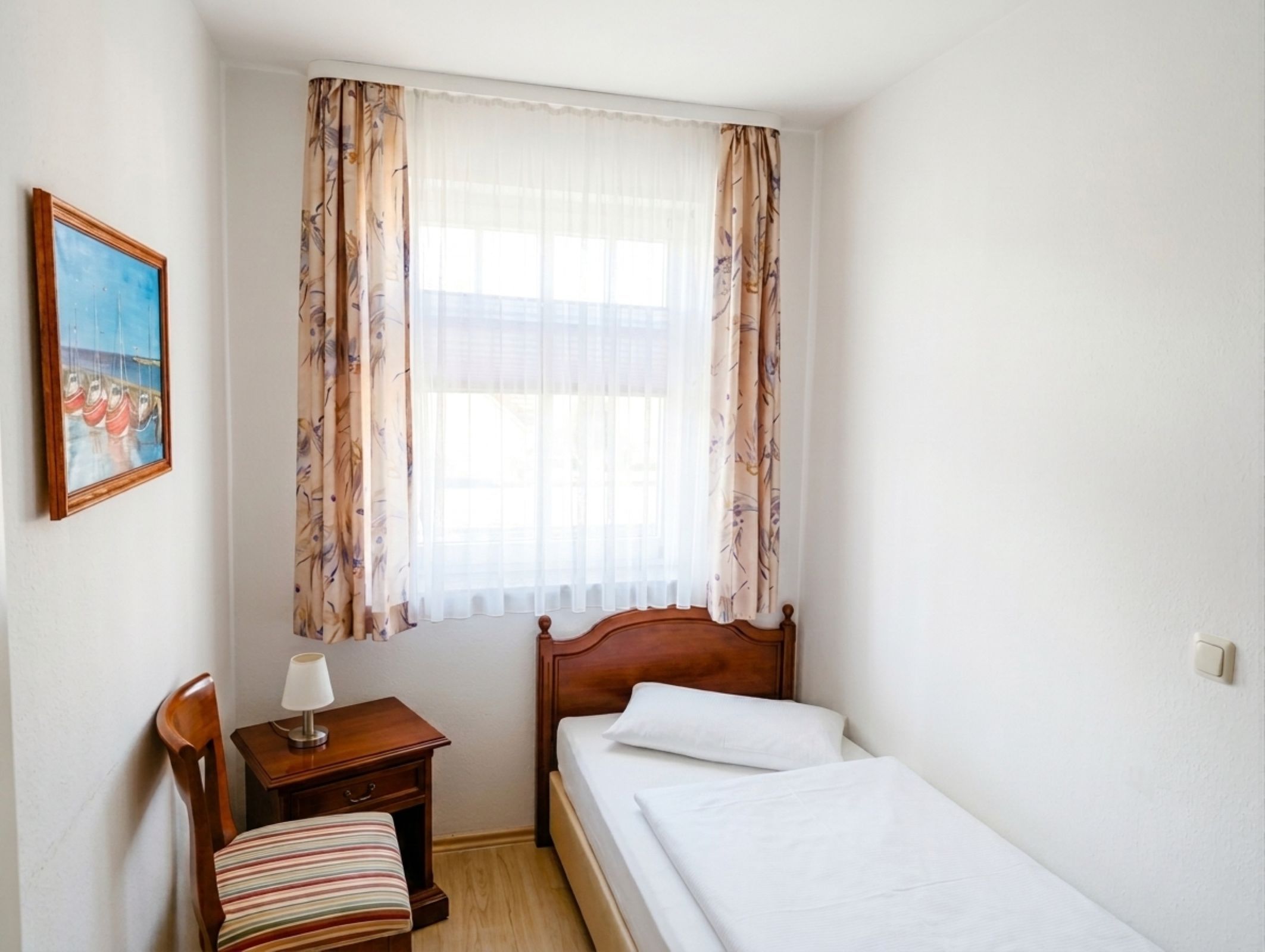 Ein Schlafzimmer mit einem Bett, einem Nachttisch, einem Sessel, einem Bild und einem Fenster mit Vorhängen.