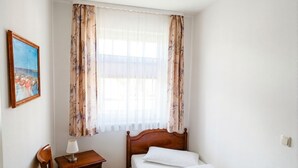 Een slaapkamer met een bed, nachtkastje, stoel, schilderij en een raam met gordijnen.