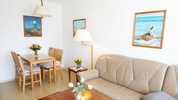 Una habitació amb taula de dinar de fusta, dues cadires, un sofà beix, una petita taula auxiliar amb una làmpada, i dos quadres emmarcades a les parets.