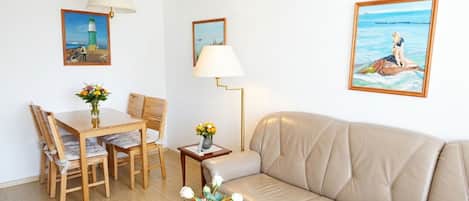 Une pièce comprenant une table à manger en bois, deux chaises, un canapé beige, une petite table d’appoint avec une lampe, et deux œuvres d’art encadrées accrochées aux murs.