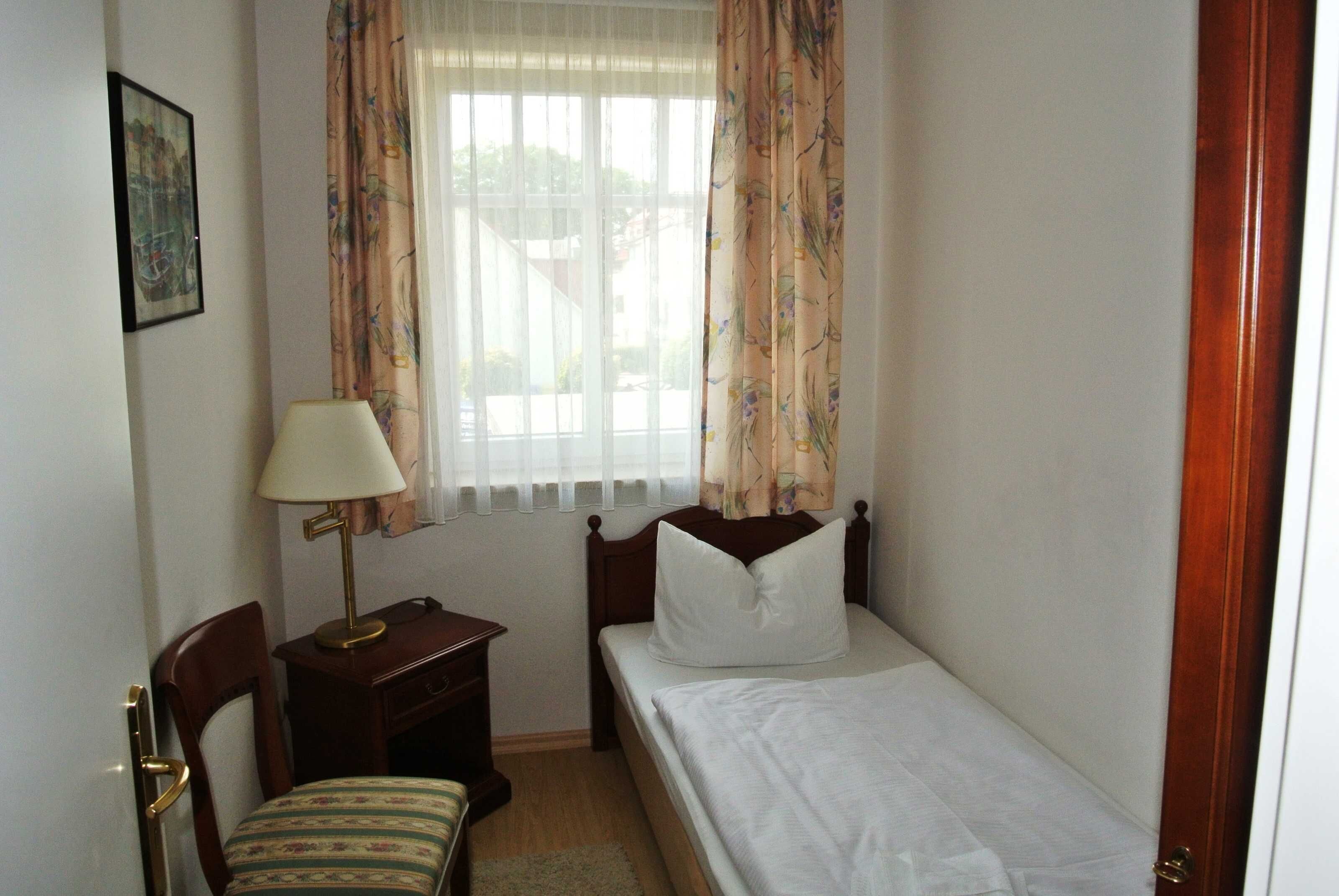 Ein kleines, aufgeräumtes Schlafzimmer mit einem Einzelbett, einem Nachttisch, einem Stuhl und einem Fenster mit Vorhängen.