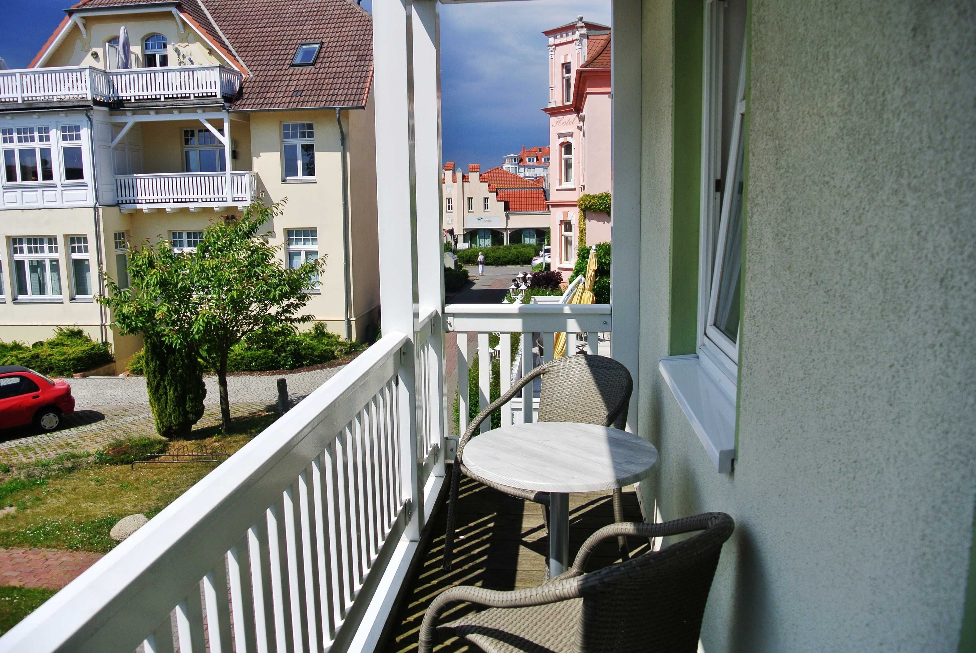 A balcony with wicker chairs and a small table, overlooking a street with buildings and a parked car.