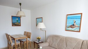A living room with a wooden table, beige sofa, and a vase with flowers. There are paintings on the walls and a lamp on the table.