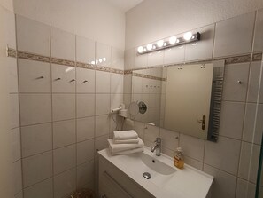 A bathroom with a white sink, mirror, and towel stack. There is a shower area with a glass door and a towel rack. - Apartment Rosenhof 04 - Wh. 04 (Kühlungsborn)