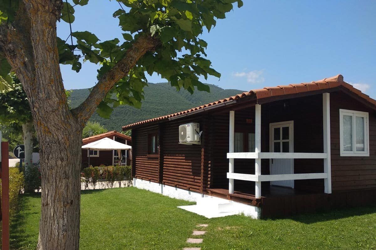 Una cabaña de madera con un porche, una estructura pequeña adosada y un árbol en primer plano.
