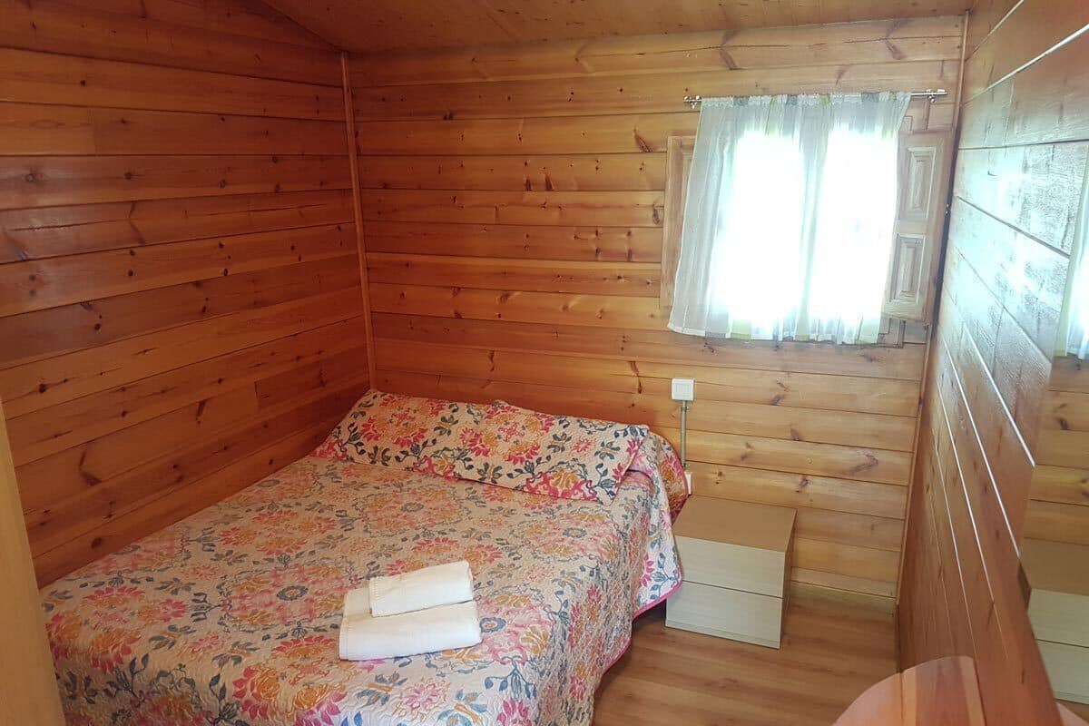 Una habitación tipo cabaña de madera con una cama, una mesita de noche pequeña y una ventana con cortinas.