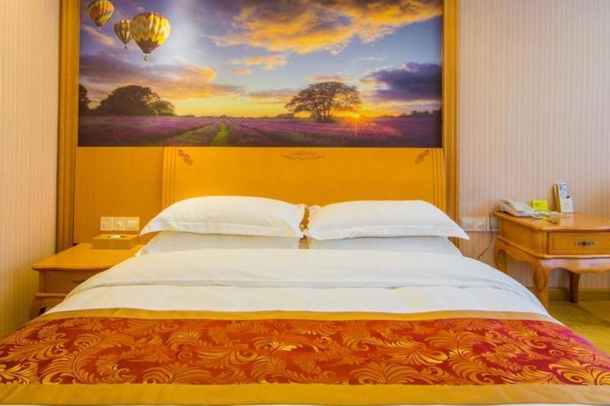 A hotel room with a large bed, a nightstand, a wooden headboard, and a painting of a sunset with hot air balloons.