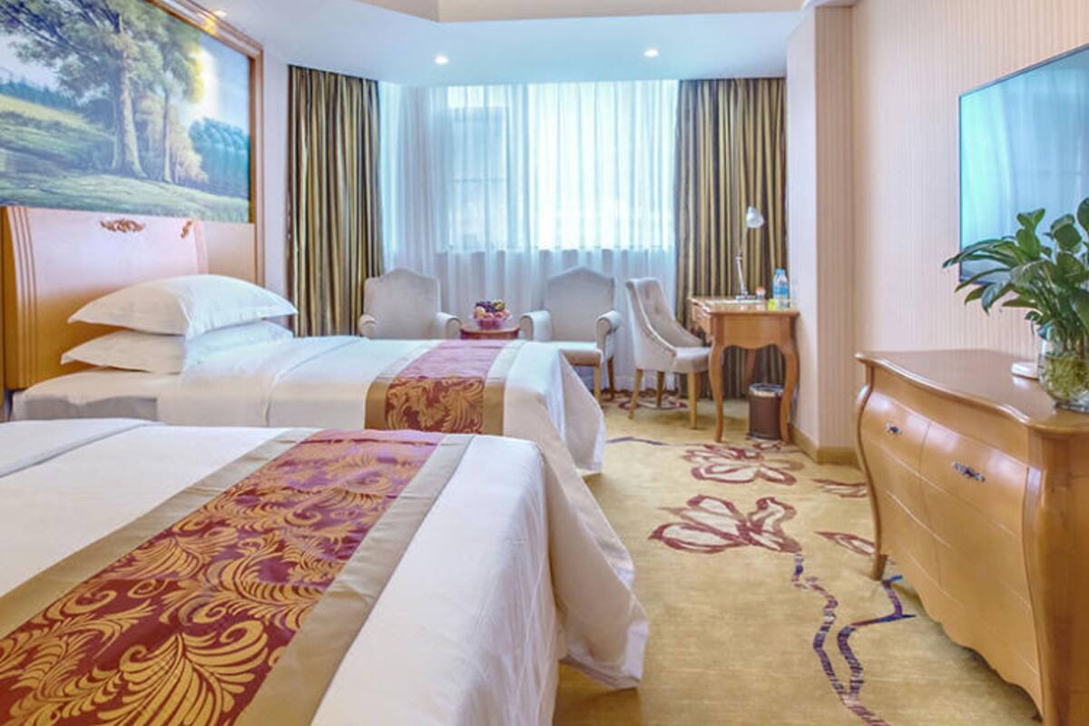 A hotel room with two beds, a desk, a chair, a TV, and a large window with curtains.