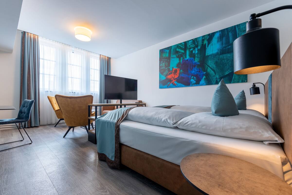 Une chambre d’hôtel moderne, équipée d’un lit, d’un bureau, d’une chaise, d’une télévision et ornée d’un tableau au mur.