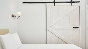 Une chambre avec un lit blanc, une lampe fixée au mur et une porte de style rustique, également blanche.