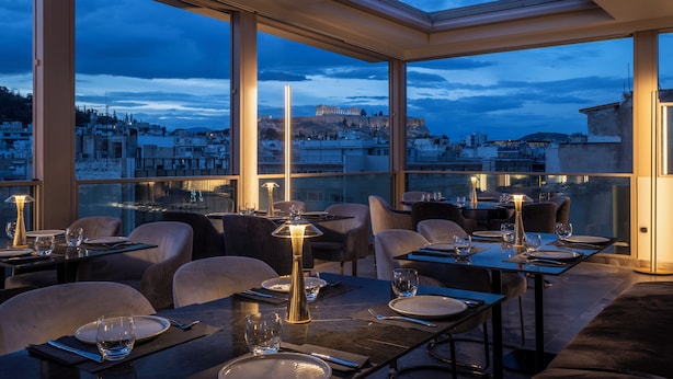 A rooftop restaurant with tables set for dining, large windows offering a view of a cityscape and a historical building, and modern interior design. - Heritage Hill (Athens)