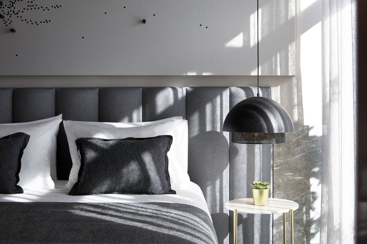 A modern bedroom with a large bed, a bedside table with a lamp, and a view of the outdoors through large windows.