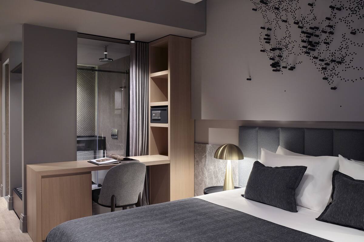 A modern hotel room with a bed, a desk, a chair, a shower, and a wall-mounted light fixture.