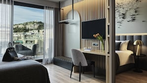 A modern hotel room with a large bed, a desk with a chair, a view of the city, and a large window with curtains. - Heritage Hill (Athens)