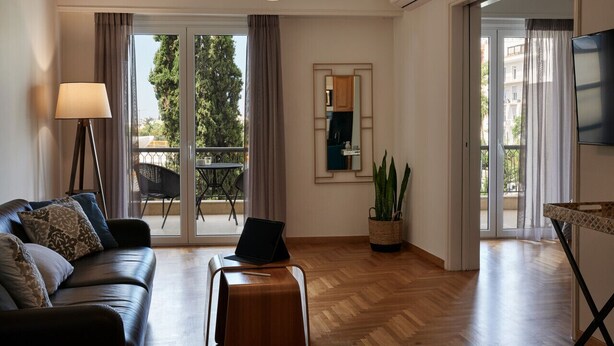 A modern living room with a sofa, a coffee table, a lamp, a laptop, and a TV. There is a sliding glass door leading to a balcony with outdoor furniture. - Chic One Bedroom Apartment with Hilton View (Athens)