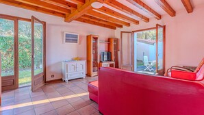 A spacious living area with a red sofa, wooden beams, a white cabinet, and a sliding glass door leading to an outdoor patio. - Charming 2-BR House with Terrace, Near Beach & Town Center, Sleeps 6 (Biscarrosse Plage)
