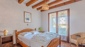 A bedroom with a wooden bed, a nightstand, a green lamp, a wicker chair, and a large window with a view. - Charming 2-BR House with Terrace, Near Beach & Town Center, Sleeps 6 (Biscarrosse Plage)