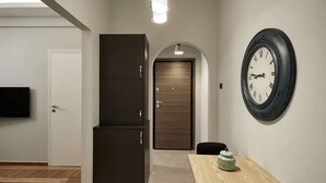 A hallway with a wooden door, a dark cabinet, a round wall clock, and a small table with a decorative item. - Charming 1bd Apt next to Concert Hall (Athina)