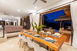 A modern dining area with a wooden table set for six, white chairs, and a view of the outdoors at dusk. - Vista Mar (Koh Samui)
