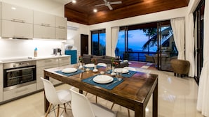 A modern kitchen with white cabinets, a wooden ceiling, and a dining area set for four with a wooden table and white chairs. - Vista Mar (Koh Samui)