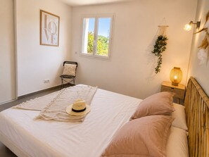 A bedroom with a bed, a window, a chair, a bedside table with a lamp, and a wall-mounted plant.