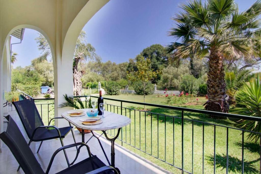 Ein Balkon mit Tisch und Stühlen, einer Flasche und einem Glas, mit Blick auf einen Garten mit Palmen und einem Giraffe.