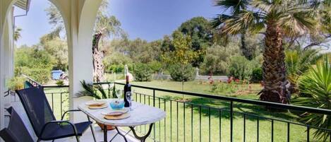 Ein Balkon mit Tisch und Stühlen, einer Flasche und einem Glas, mit Blick auf einen Garten mit Palmen und einem Giraffe.