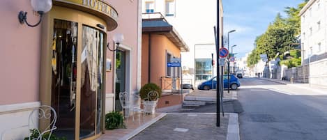 Un hotel con entrada de vidrio, asientos exteriores y un coche azul estacionado en la calle.