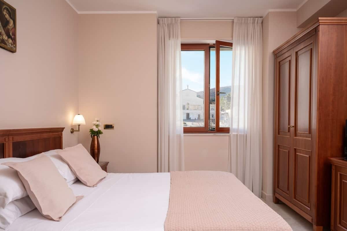 Ein Hotelzimmer mit Bett, Nachttisch, Lampe, einem Fenster mit Vorhängen und einem Schrank.