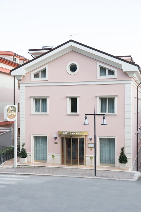 A pink building with a sign reading "Hotel San Pio." It has a modern facade, large windows, and a small entrance with a door. - Hotel San Pio (San Giovanni Rotondo)