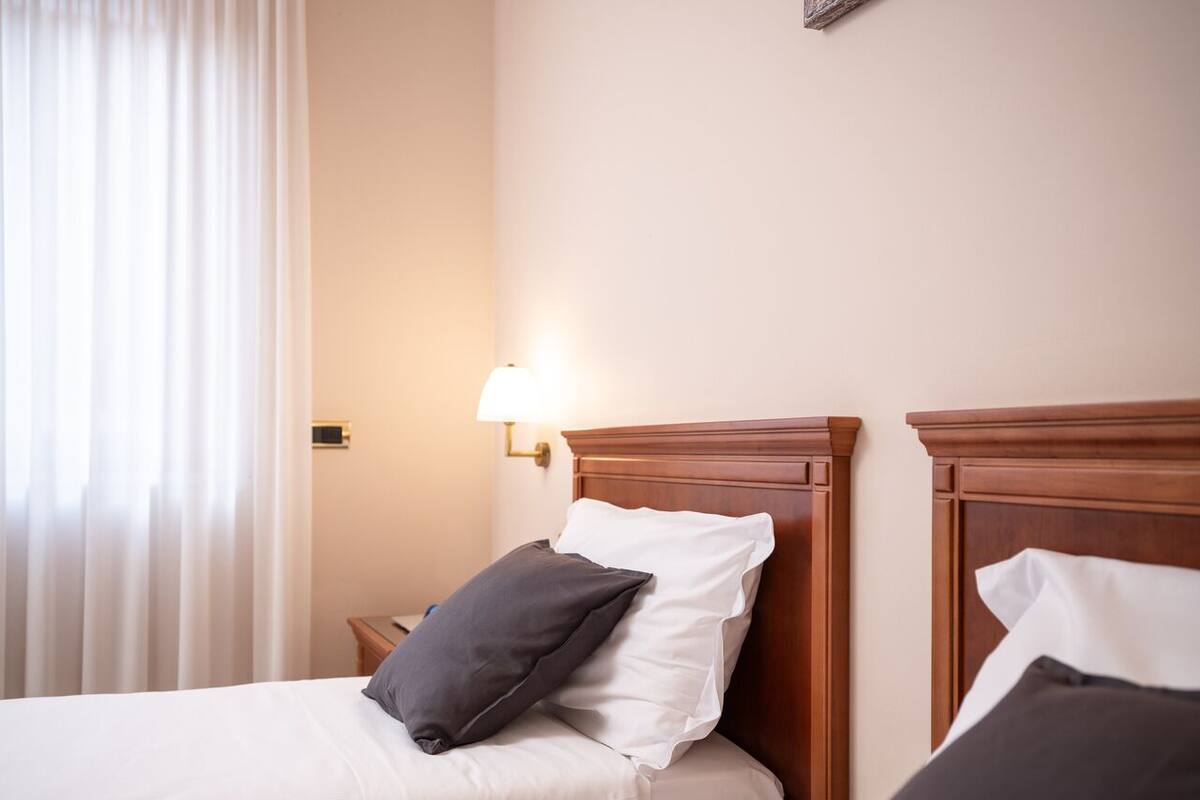 Ein Hotelzimmer mit zwei Betten, jeweils mit einem hölzernen Kopfteil, weißer Bettwäsche und einem einzelnen dunkel gefärbten Kopfkissen. Eine Wandleuchte ist auf der linken Seite sichtbar.