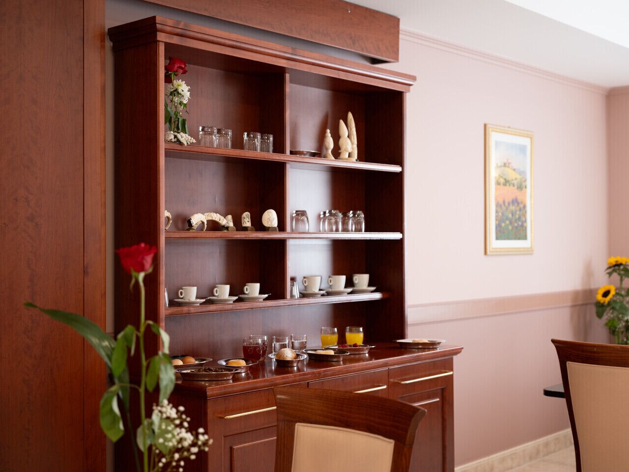 A wooden buffet with glassware and dishes, a vase with a red rose, and a painting on the wall.