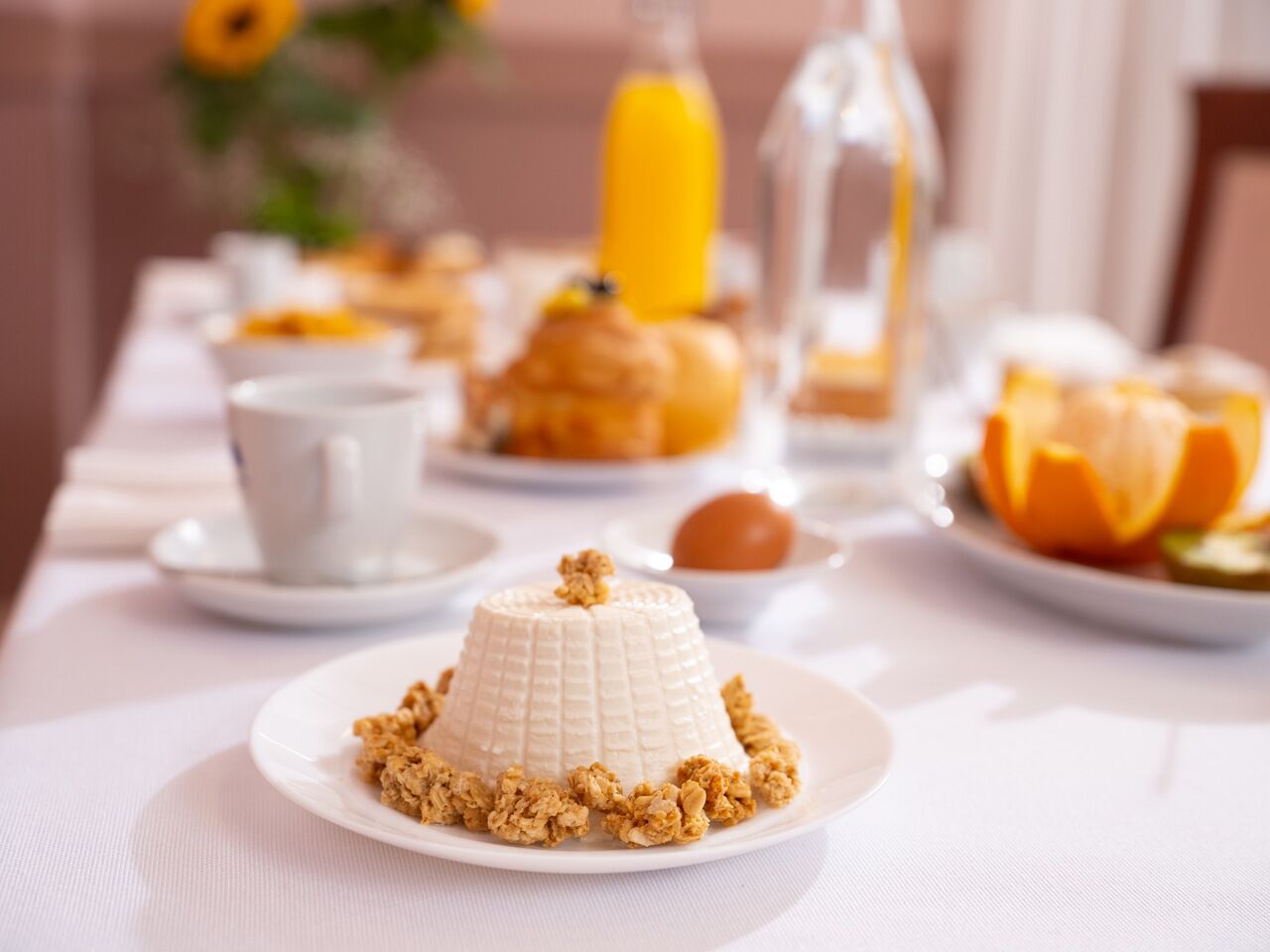 Colazione con un dessert panna cotta su un piatto, accompagnata da granola, un bicchiere di succo d'arancia e una tazza.