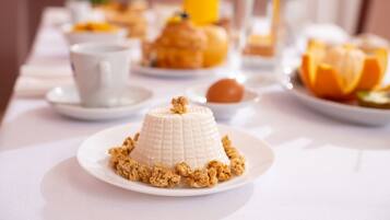 Colazione con un dessert panna cotta su un piatto, accompagnata da granola, un bicchiere di succo d'arancia e una tazza.