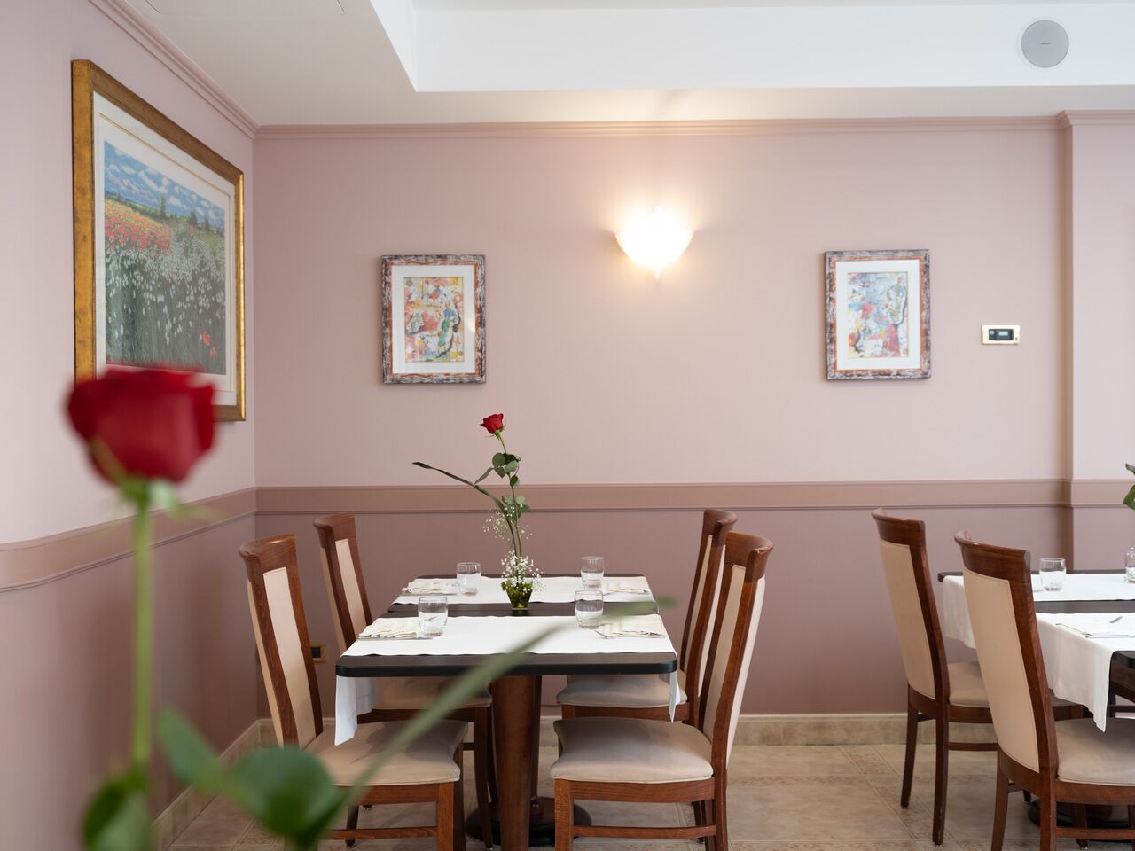 Een eetruimte met een tafel voor vier personen, omringd door houten stoelen. Twee ingelijste kunstwerken hangen aan de muren, er ligt een roos op de tafel en een lamparmatuur hangt aan het plafond.