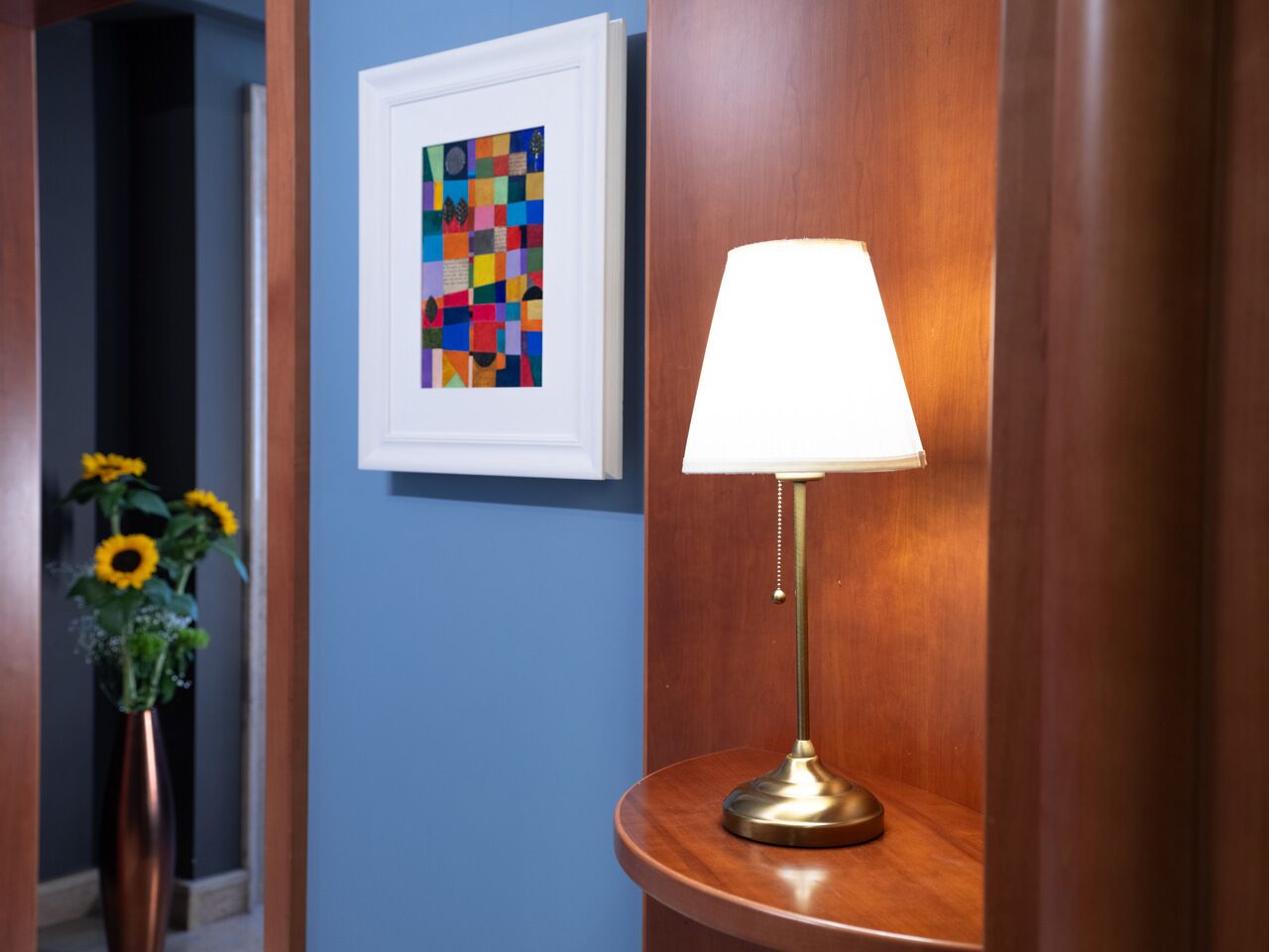 A hallway with a framed abstract painting, a round table with a lamp, and a vase with sunflowers.
