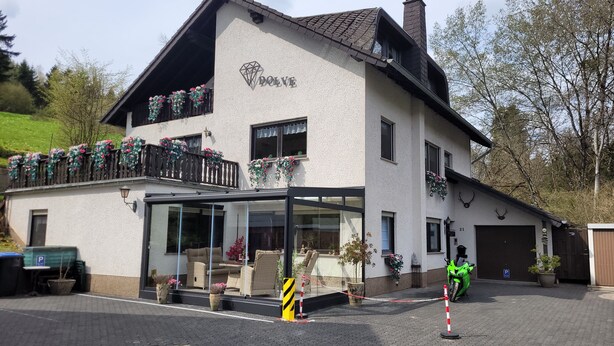 A two-story building with a balcony adorned with flowers, a glass entrance, and a parking area with a green car. - Familiehuis Dolve (Muerlenbach)