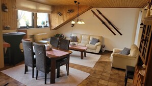 A cozy living room with a wooden ceiling, a sofa set, a dining table with chairs, and a kitchen area with a window. - Familiehuis Dolve (Muerlenbach)