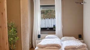 A bedroom with two beds, a window with curtains, a bedside table with a lamp, and a potted plant. - Familiehuis Dolve (Muerlenbach)