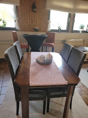A dining table with a patterned tablecloth, surrounded by wicker chairs, in a room with wooden walls and a window with curtains.