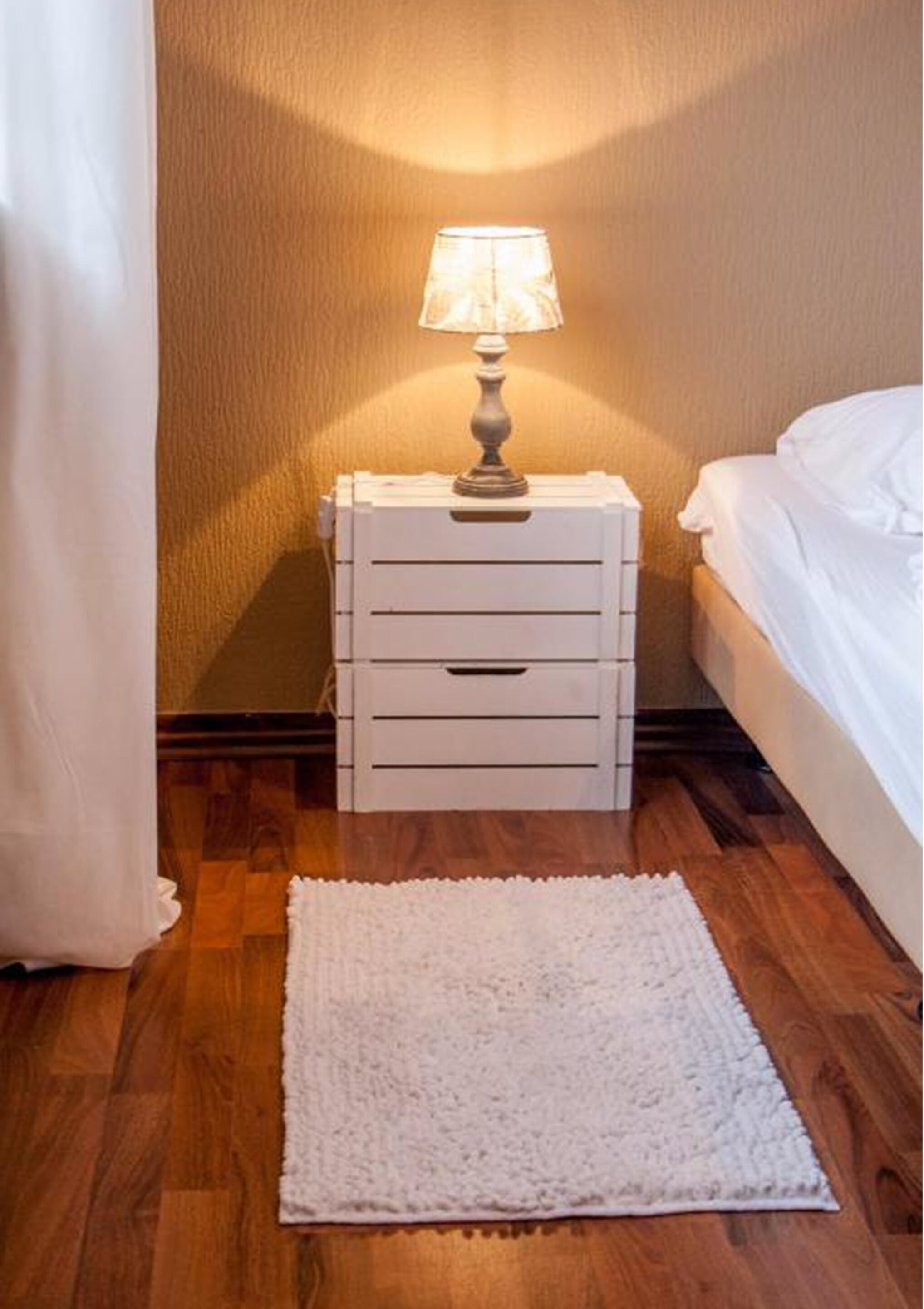 Un letto rifatto con copriletto bianco, una lampada da comodino su un comodino bianco e un tappeto bianco su un pavimento in legno.
