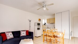 A compact living space with a dark blue sofa, a small white table, a wooden dining table with chairs, a ceiling fan, and a kitchenette with white cabinets and appliances. - Biscarrosse: Appartement T3, 2 Chambres, Terrasse, Parking Privé à 50m de la Plage (Biscarrosse Plage)