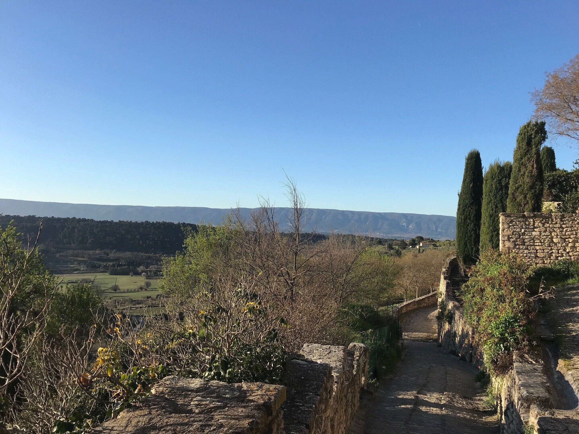 Ein steiniger Weg führt durch eine ländliche Gegend mit freiem Blick auf weit entfernte Hügel und eine Steinsäule auf der rechten Seite.