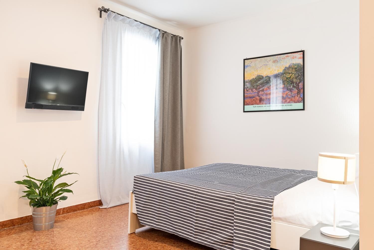 A hotel room with a bed, a TV, a painting, a plant, and a lamp.