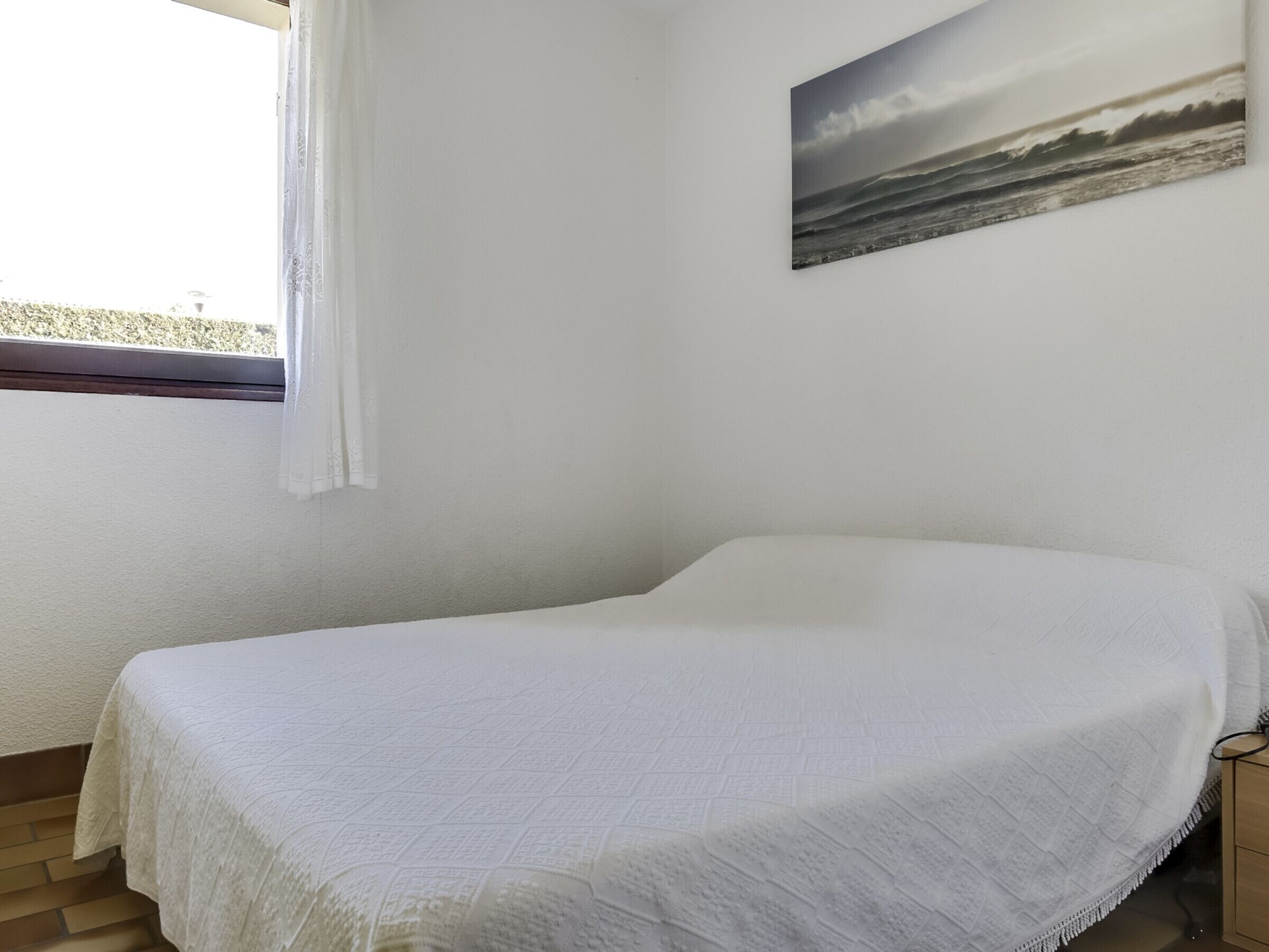A single bed in a room with a plain white wall, a window with a white curtain, and a painting of a seascape.