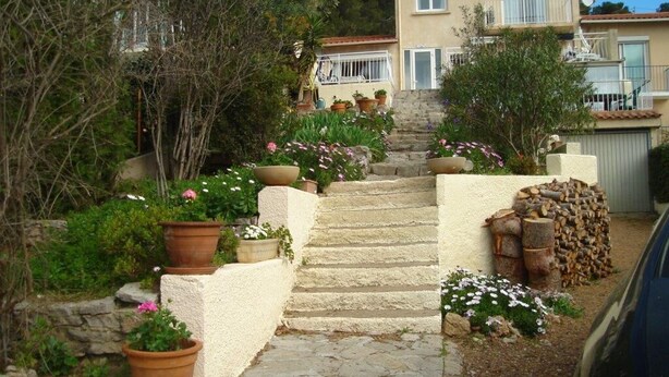 Una escalera de piedra conduce a una casa de fachada color beige, rodeada de macetas con plantas y flores.