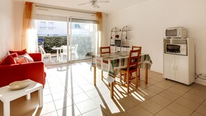 Un salon lumineux avec un canapé rouge, une table à manger avec des chaises, un ventilateur de plafond et un coin cuisine équipé d’un four à micro-ondes et d’un four traditionnel.