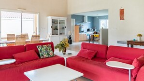 Amplia sala de estar con sofás rojos, una mesa de centro blanca y un comedor con sillas de mimbre. Se ve una cocina al fondo con electrodomésticos modernos.
