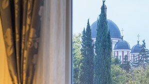 Una ventana con vista a un edificio con cúpulas y cruces, cortinas y una cama con una colcha roja estampada con flores.