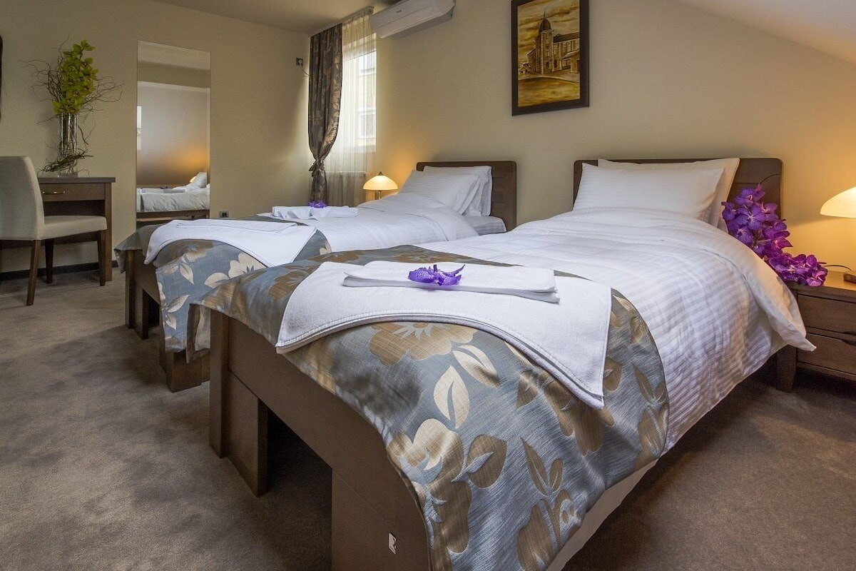 Quarto de hotel com duas camas, uma escrivaninha, uma cadeira, uma mesinha, uma luminária e um claraboia.